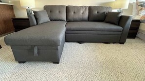 A grey sectional sofa with a matching ottoman in a room with a carpeted floor and two bedside tables with lamps. - Studio w/ King Bed, Ocean Views, Pet Friendly! (Lincoln City)