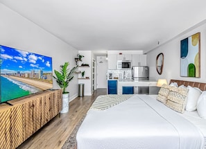 A modern hotel room with a large bed, a TV, a kitchenette, and a view of the beach.
