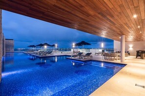 A rooftop infinity pool with lounge chairs and umbrellas, surrounded by a wooden deck and a view of the city skyline. - Stunning Rooftop Views - Steps from the Sand (Playa del Carmen)