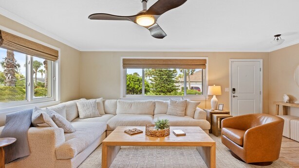 Ein Wohnzimmer mit Ecksofa, Couchtisch, Deckenventilator und großen Fenstern mit Blick auf Grünflächen.
