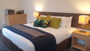 Een netjes opgemaakt bed met een donkerblauwe deken, een houten nachtkastje met een lamp, en een magnetron op een dressoir.
