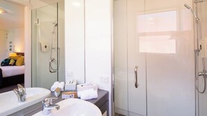 Een moderne badkamer met twee wastafels, een glazen douchecabine en een zichtbaar bed in de aangrenzende kamer.