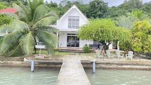 A white house with a large window, surrounded by lush greenery and a small dock leading to a body of water. - Peaceful house by the sea (Îles Sous-le-Vent)