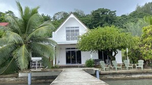 A white building with a balcony, a concrete pathway leading to the entrance, and a palm tree to the left. - Peaceful house by the sea (Îles Sous-le-Vent)