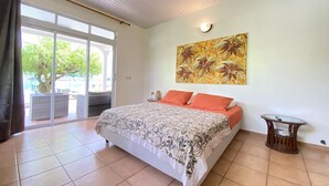A bedroom with a bed, a painting, a small table, and a view through a sliding door. - Peaceful house by the sea (Îles Sous-le-Vent)