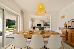 A dining area with a wooden table, white chairs, woven placemats, and a hanging light fixture.