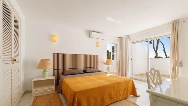A hotel room with a bed, two bedside tables, a lamp, a wall-mounted air conditioner, and a view of the outdoors.