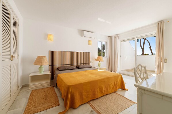 A hotel room with a bed, two bedside tables, a lamp, a wall-mounted air conditioner, and a view of the outdoors.