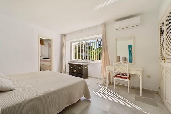 A bedroom with a bed, a dresser, a desk with a chair, a window with curtains, and a mirror on the wall.