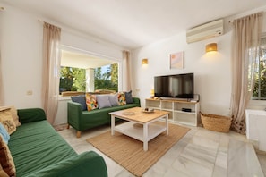 A living room with a green sofa, a coffee table, a flat-screen TV, and a large window with curtains.