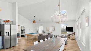 A spacious living area with a wooden dining table, white chairs, a pool table, and a modern kitchen with stainless steel appliances.