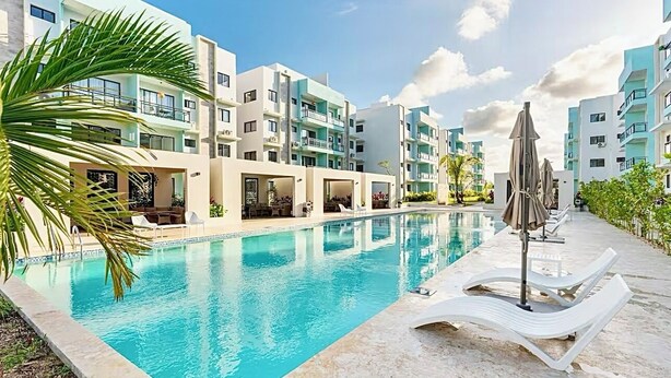A swimming pool with lounge chairs and umbrellas, surrounded by multi-story buildings with balconies.