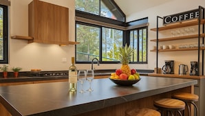 Una cocina moderna con una isla central, taburetes de bar, una estación de café y vistas a árboles a través de amplios ventanales.