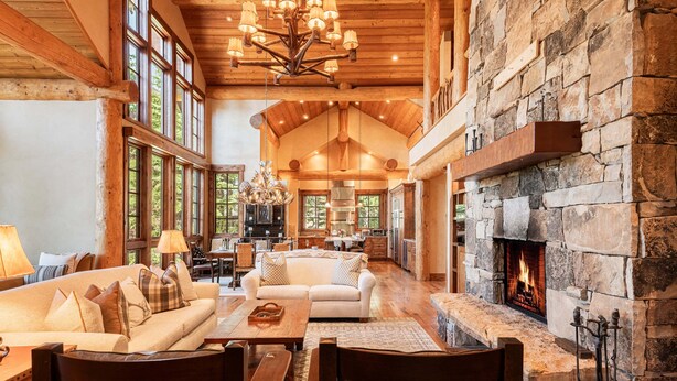 A spacious living room with a stone fireplace, wooden ceiling beams, and large windows.
