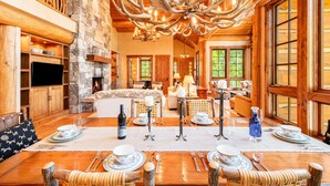 A spacious interior with a large wooden dining table set for a meal, a stone fireplace, and a unique antler chandelier.