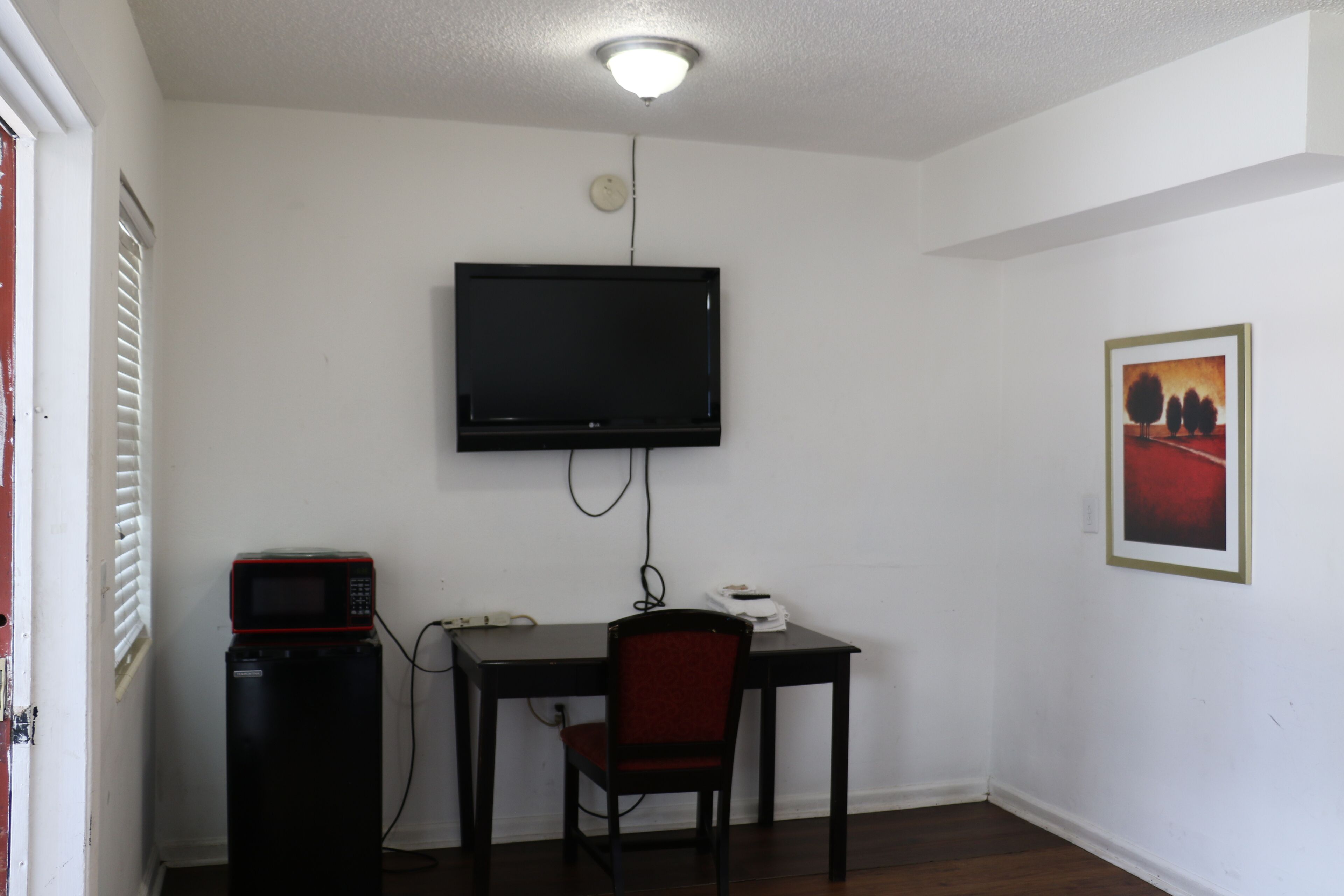 Habitación con televisión de pantalla plana montada en la pared, un escritorio con una silla, microondas y un cuadro enmarcado.