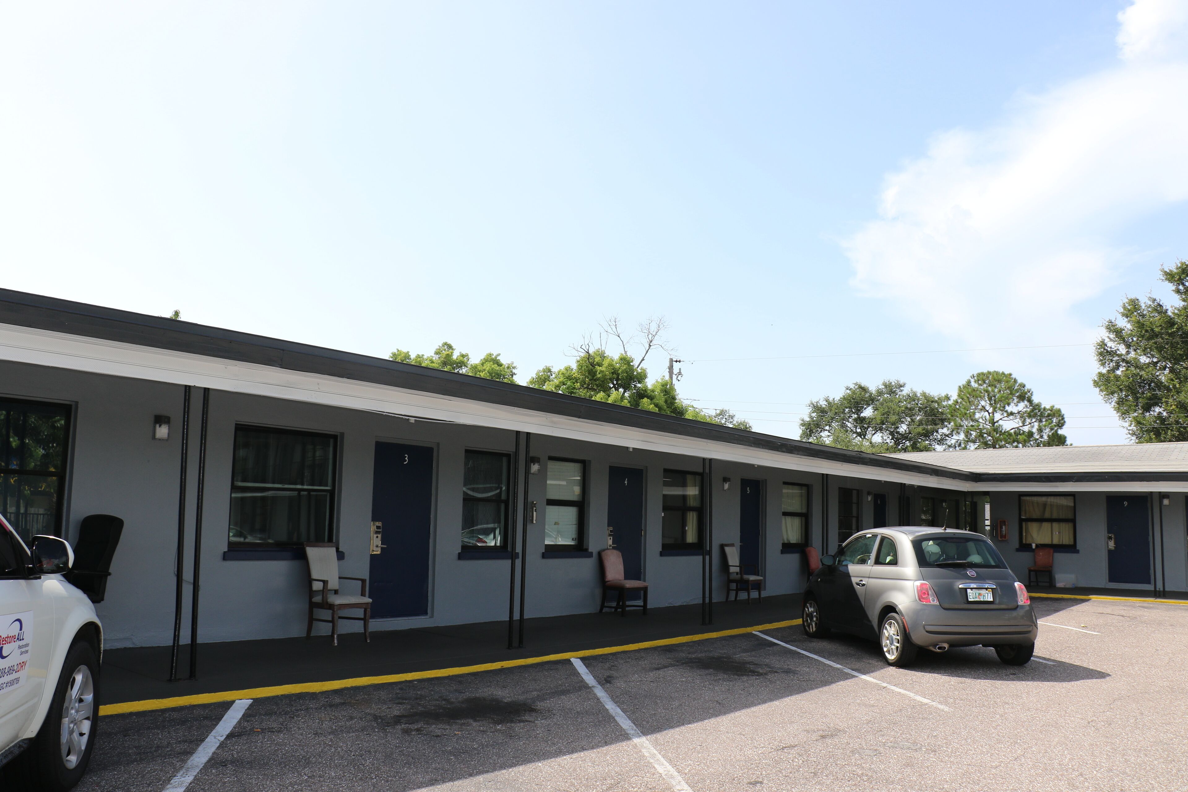 Un motel con una fila de habitaciones, cada una con puerta y ventana. Se observa un estacionamiento con un coche estacionado al frente.