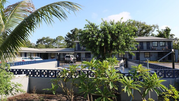 Un motel con fachada azul y blanca, una zona de estacionamiento techada y una palmera en primer plano.