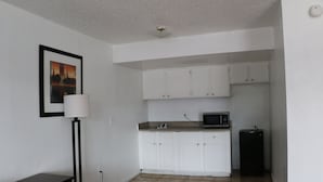 Una cocina pequeña y limpia con armarios blancos, un microondas y un refrigerador. Hay un cuadro en la pared y una lámpara de pie cerca de la cama.