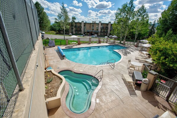 Une piscine avec un jacuzzi, entourée de chaises longues et de parasols, avec un bâtiment en arrière-plan.