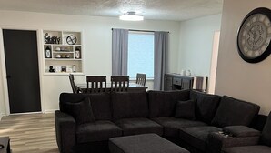 A living room with a sectional sofa, ottoman, and a rug. There is a dining table with chairs, a wall clock, and a window with curtains. - Charming 3-bedroom family friendly home in York (York)