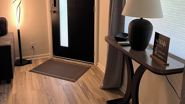 A hotel room with a wooden floor, a standing lamp, a table with a lamp and a sign, a door, and a window with curtains. - Charming 3-bedroom family friendly home in York (York)