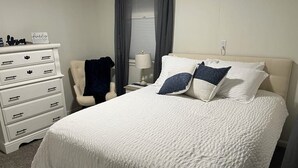 A neatly made bed with white bedding and two pillows, a white dresser with drawers, a chair, and a lamp on a bedside table. - Charming 3-bedroom family friendly home in York (York)