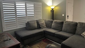 Una sala de estar con un sofá seccional, una lámpara y una ventana con persianas.