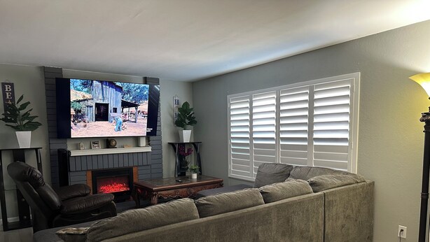 Una sala de estar con un sofá seccional, una chimenea, un televisor montado en la pared y un ventanal con persianas.