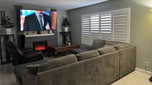 Una sala de estar con una televisión de pantalla plana grande, chimenea, sofá y ventana con persianas.