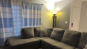 Un sofá modular con un plaid estampado, una lámpara y una cortina escocesa en una habitación con una puerta y un interruptor de luz en la pared.