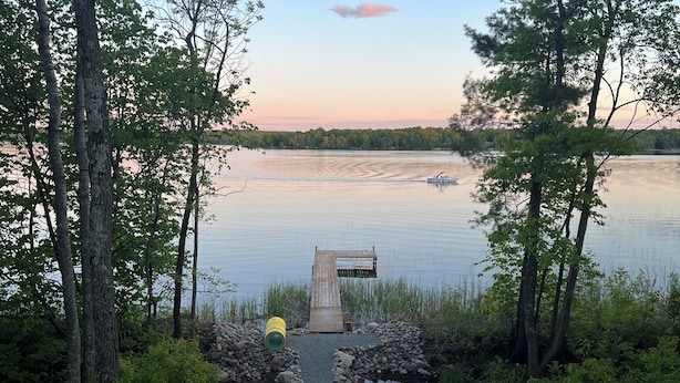 A gravel area with Adirondack chairs and a fire pit, a yellow kayak, and a wooden dock extending into a lake surrounded by trees.