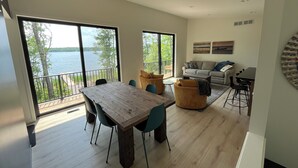 A spacious living area with a large wooden table, comfortable seating, and a balcony overlooking a lake.