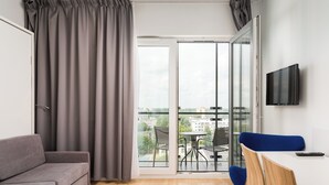Ein modernes Zimmer mit grauem Sofa, einem kleinen Tisch mit Stühlen, einem an der Wand befestigten Fernseher und einer Glastür, die auf einen Balkon mit Gartenmöbeln führt.
