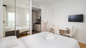 A hotel room with a bed, a desk, a chair, a TV, and a kitchenette in the background.