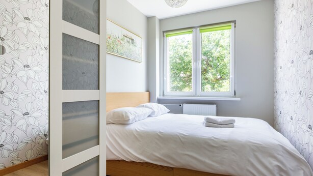 A bedroom with a bed, a radiator, a window with green blinds, and a wall with floral wallpaper. - Lecha Comfort Apartment (Gdańsk)