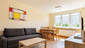 A modern living room with a grey sofa, wooden dining table, and chairs. A colorful abstract painting is on the wall, and there is a window with a view of buildings. - Lecha Comfort Apartment (Gdańsk)