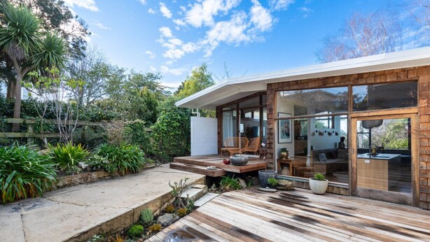 A modern house with a wooden deck, outdoor seating, and a glass door leading to a kitchen area.