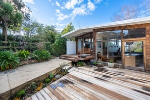 Een modern huis met een houten terras, zitplaatsen buiten en een glazen deur die leidt naar een keuken.