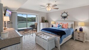 A bedroom with a bed, a ceiling fan, a nightstand, a chair, a small table, a lamp, a window with a view of greenery, and a wall-mounted mirror.