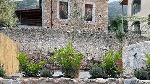 Un mur de pierre avec, en arrière-plan, un bâtiment au style rustique, doté d’un balcon et d’une fenêtre avec des volets.