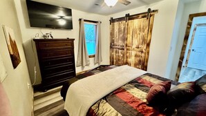 A bedroom with a bed, a dresser, a TV, a ceiling fan, and a window with curtains. - Drew’s Delight - Relax in Comfort!  Modern, Fully Remodeled, No Stairs! (Mountain Grove)