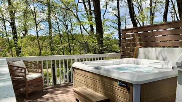 Un jacuzzi en una terraza de madera con una silla de mimbre y un banco, rodeado de árboles.