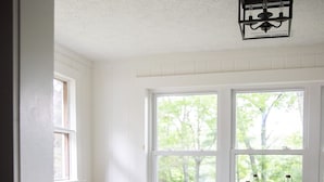Un comedor con una mesa redonda, dos sillas, un mantón de piel blanca, una cesta de latón con follaje y una ventana con vista a vegetación.