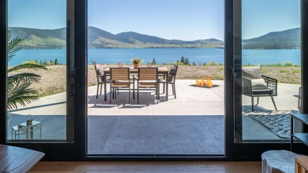 A room with a large sliding glass door leading to an outdoor patio with a table and chairs, a fire pit, and a view of a lake and mountains. - Cozy, Tranquil, & Stunning Lake View Home with Hot Tub, Sauna, and Outdoor Fun! (Elmo)