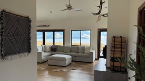 A spacious living room with a large sectional sofa, a ceiling fan, and a view of a lake through large windows. - Cozy, Tranquil, & Stunning Lake View Home with Hot Tub, Sauna, and Outdoor Fun! (Elmo)