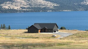 A large, dark-colored house with a sloped roof, situated on a grassy hill overlooking a large body of water. - Cozy, Tranquil, & Stunning Lake View Home with Hot Tub, Sauna, and Outdoor Fun! (Elmo)