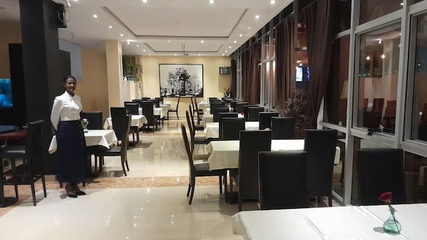 En hotellrestaurang med bord och stolar uppställda i rader, en servitör som står vid ingången och ett stort fönster på höger sida.