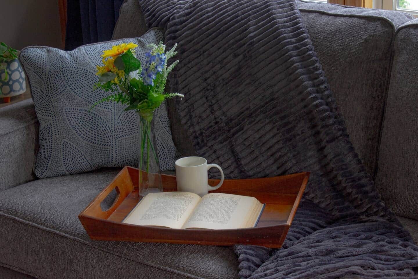Rincón acogedor para leer con un libro, una taza y un florero sobre un sofá gris.