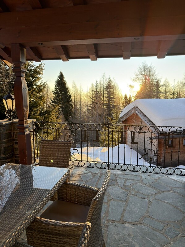 A covered outdoor seating area with stone flooring, wicker furniture, and a metal railing overlooking a snowy landscape and forest.